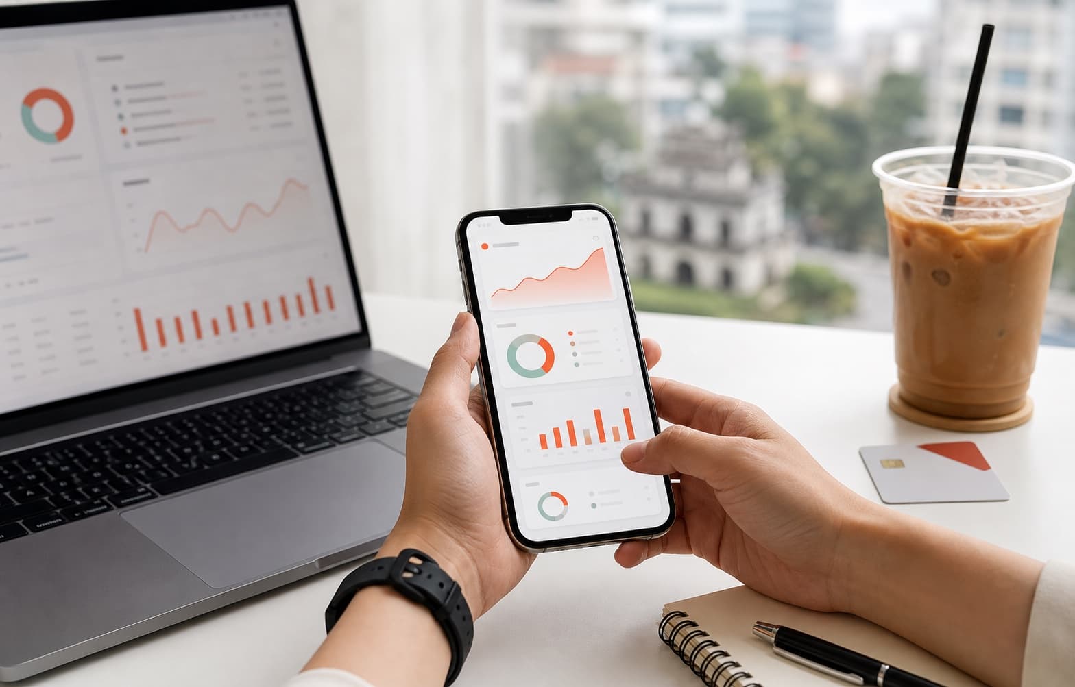 Bàn làm việc sáng với điện thoại hiển thị dashboard doanh thu, laptop phân tích chiến dịch và cà phê Việt Nam.